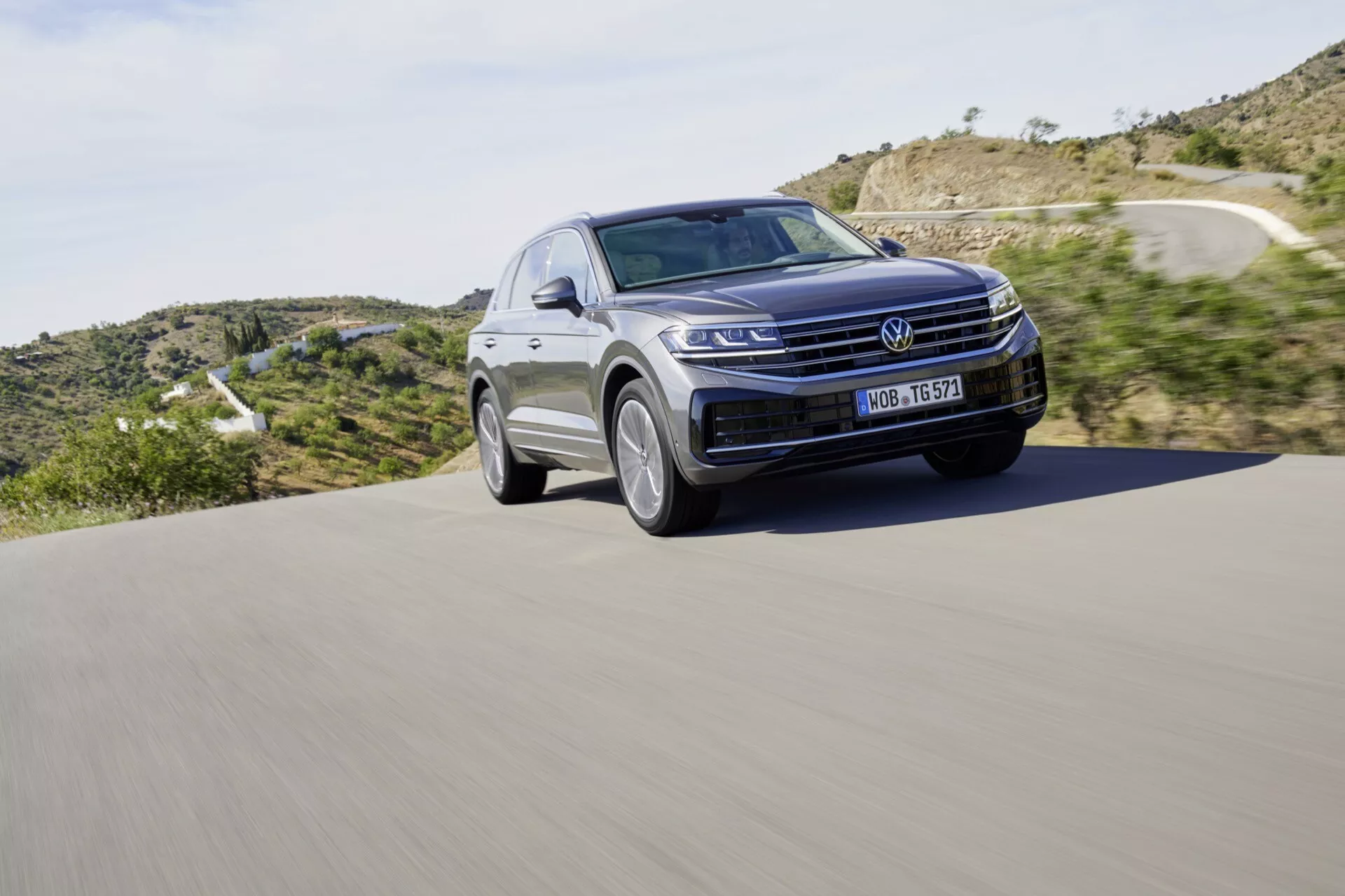 Volkswagen Touareg III Facelift SUV z roku 2023 v pohybu na asfaltové silnici, s výraznou přední mřížkou, LED světlomety a elegantní linií kapoty na pozadí přírodní krajiny, zdůrazňující moderní design a automobilovou technologii.