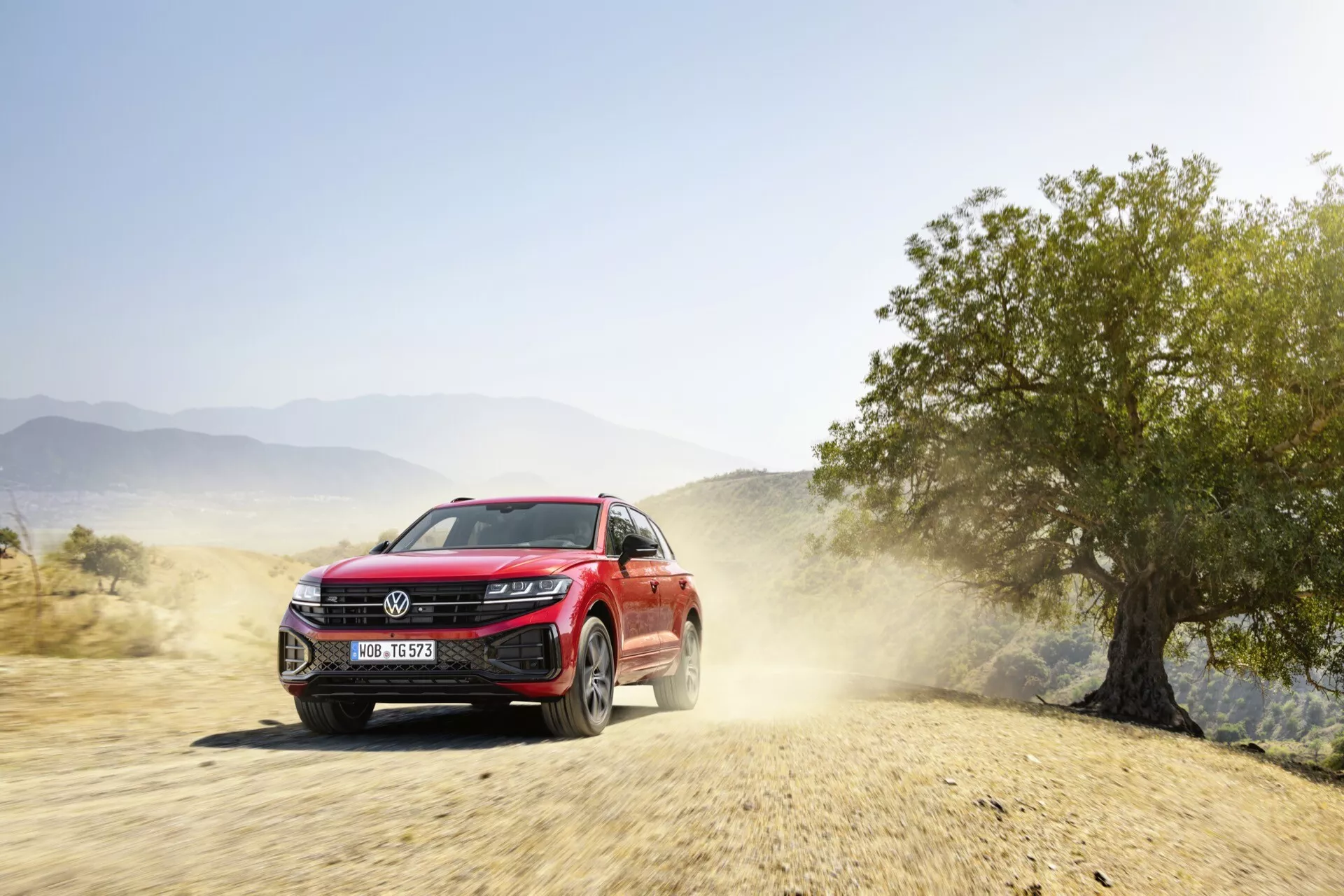 Červené SUV VW Touareg 2023 v dynamickém pohybu na nezpevněné cestě se stopou prachu, předvádějící přední fasádu, LED světlomety, mřížku s potenciálními sensory pro asistenční systémy řidiče a slitinová kola, naznačující schopnosti v terénu a moderní technologické funkce