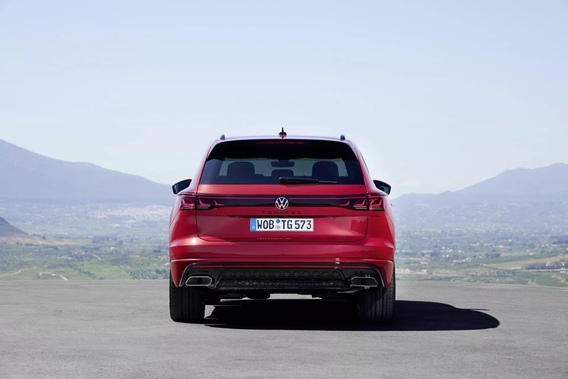 Zadní pohled na Volkswagen Touareg 2023 ukazuje celou šířku, moderní zadní světla, zadní difuzor a dvojité výfukové tipy, pod přirozeným denním světlem s čistou oblohou a kopcovitým pozadím krajiny