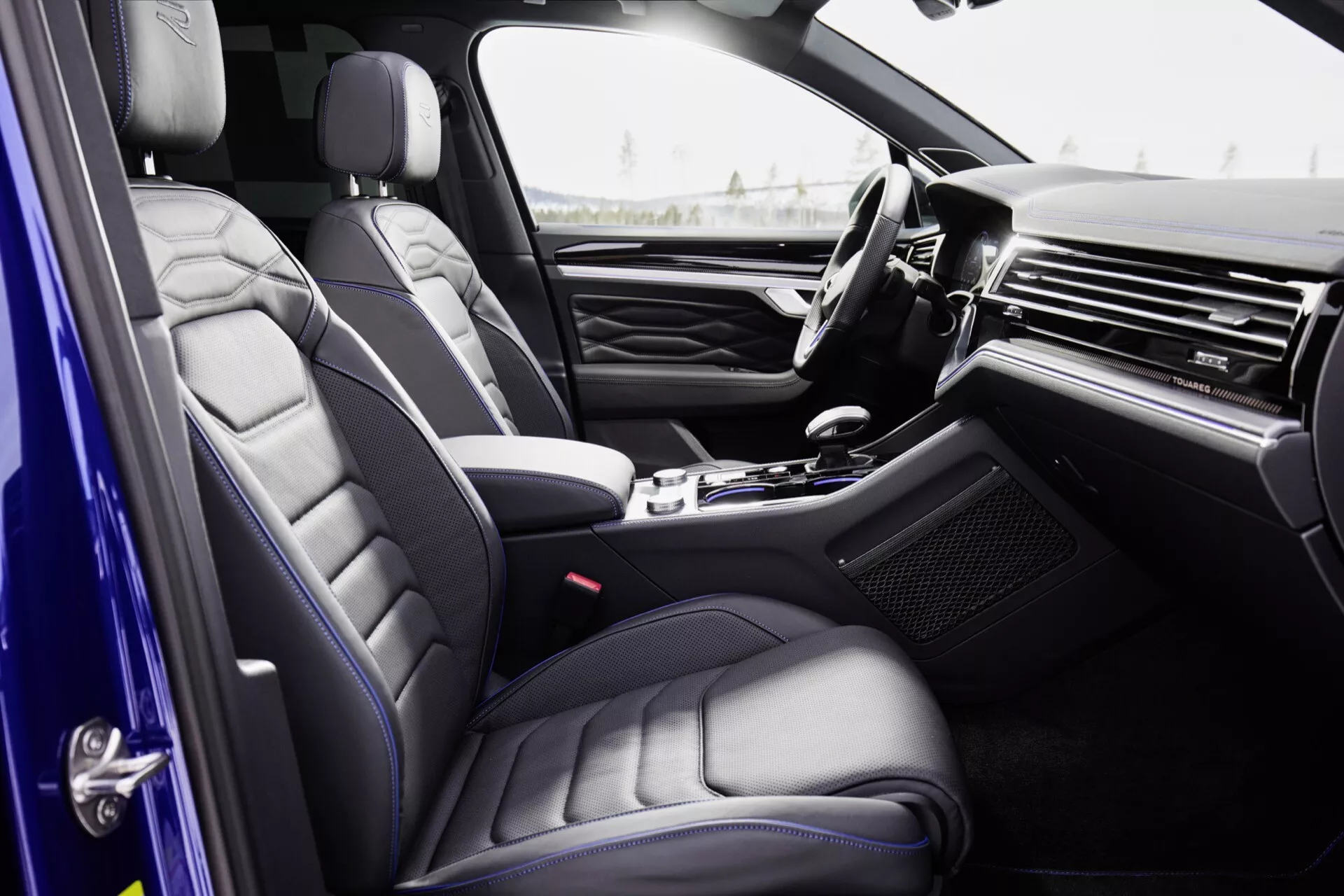 Interiér Volkswagen Touareg 2023 (III facelift) z pohledu pasažéra, zdůrazňující sedadla z prémiové kůže s prošívaným designem, elegantní palubní desku s kovovými nebo dřevěnými prvky, luxusní středovou konzolu s voličem automatické převodovky, dveřní panel s vysokou úrovní výbavy a částečně viditelný volant s integrovanými ovladači.