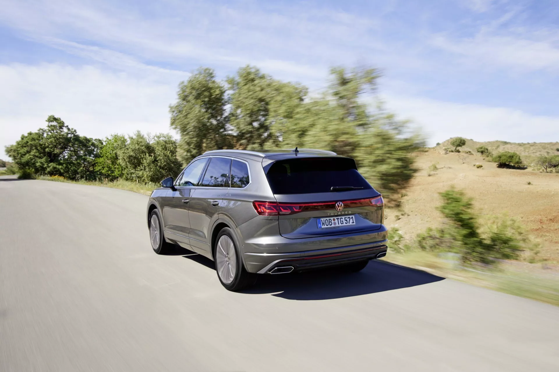 Facelift VW Touareg III z roku 2023 zachycený z pohledu ze zadního tříčtvrtečního úhlu při pohybu během dne za jasného počasí, předvádějící LED zadní světla, elegantní design kombi s mírně svažující se střechou a spoiler na střeše, částečný pohled na design slitinového kola a metalický lak.