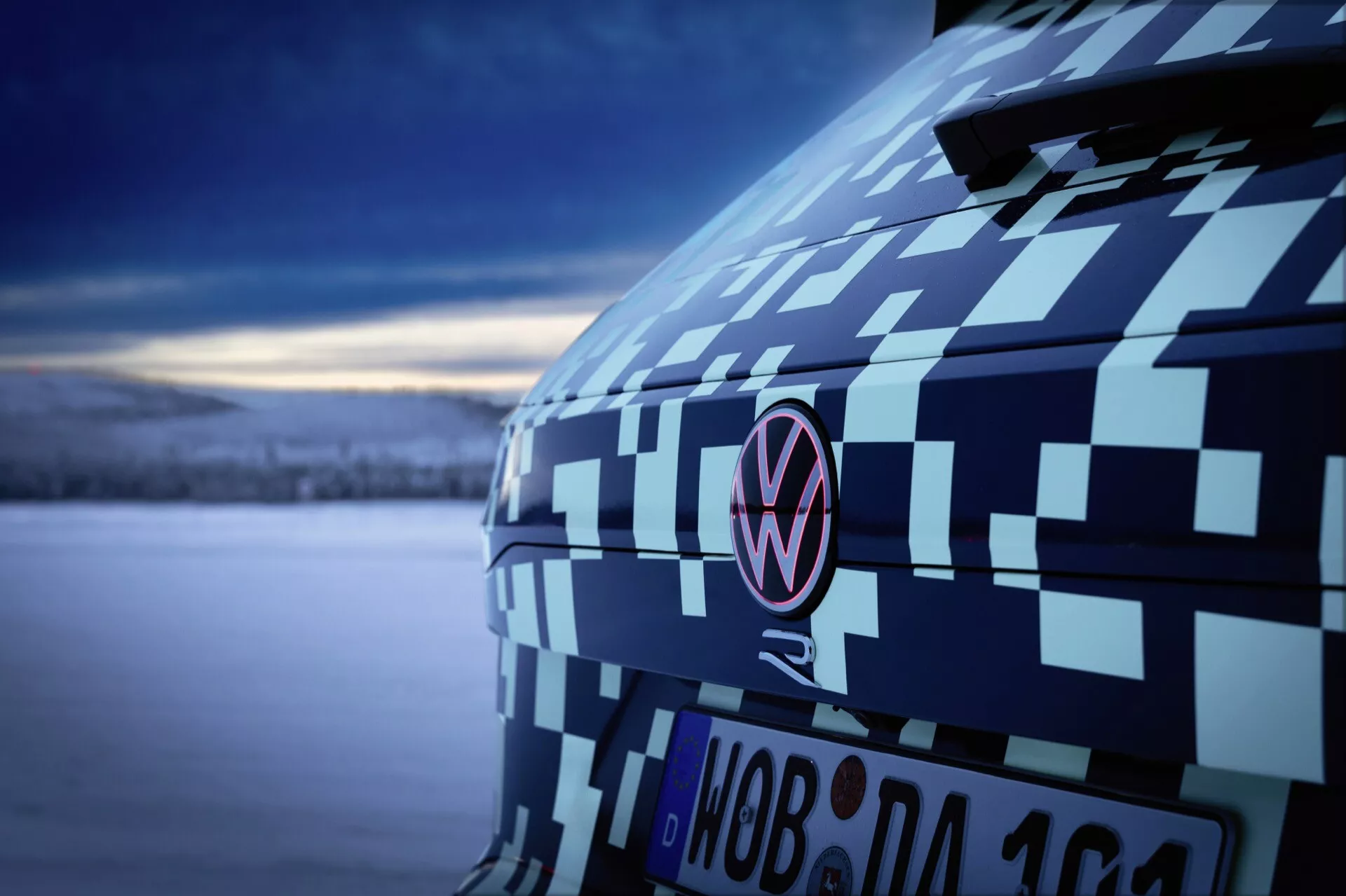 Detailní pohled na zadní část Volkswagenu Touareg (III Facelift) rok 2023 s maskovacím potahem zdůrazňujícím C-sloupek a emblém za soumraku, prezentující testování prototypů automobilů a design za měkkého, chladného osvětlení