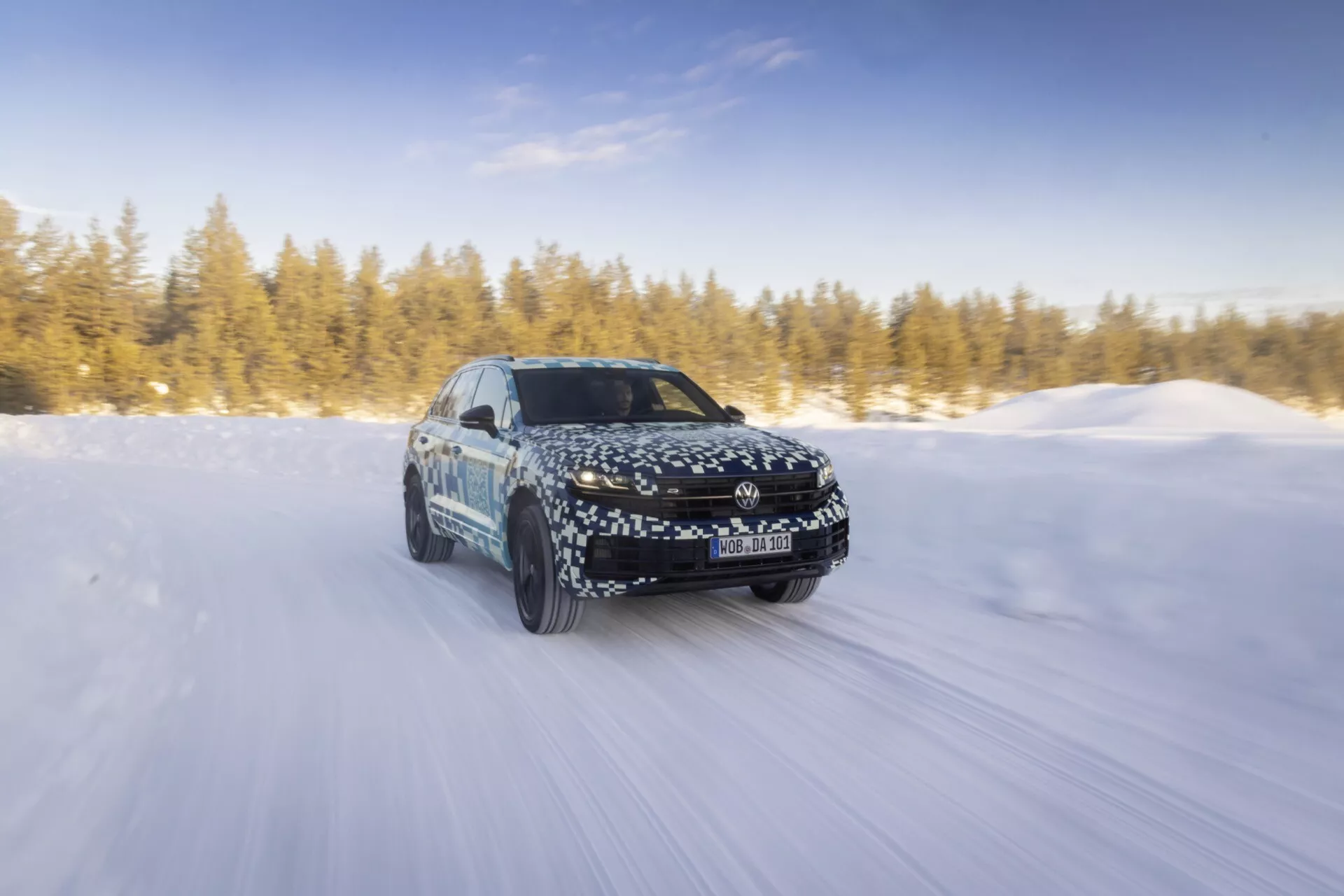 Model Volkswagen Touareg III facelift z roku 2023 dynamicky jedoucí po pokryté sněhem silnici na slunci, ukazující pohyb vpřed, LED světlomety, slitinová kola a svůj aerodynamický design s maskovacím obalem.