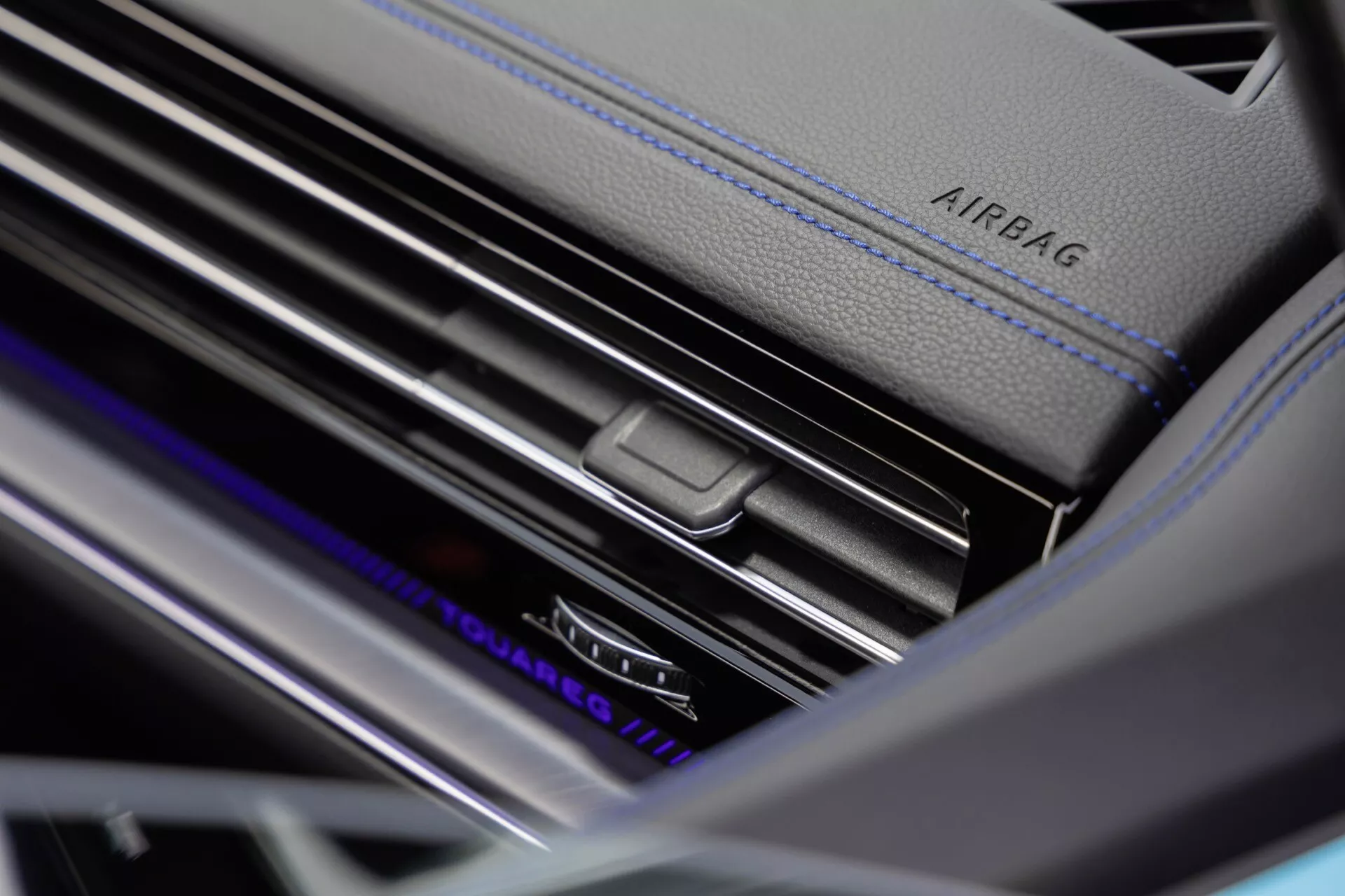 Detailní záběr interiéru VW Touareg 2023 (III-facelift) ukazující detail vzduchového ventilu na palubní desce s ovládáním klimatizace pro dvě zóny, kontrastní modré prošívání a ambientní osvětlení