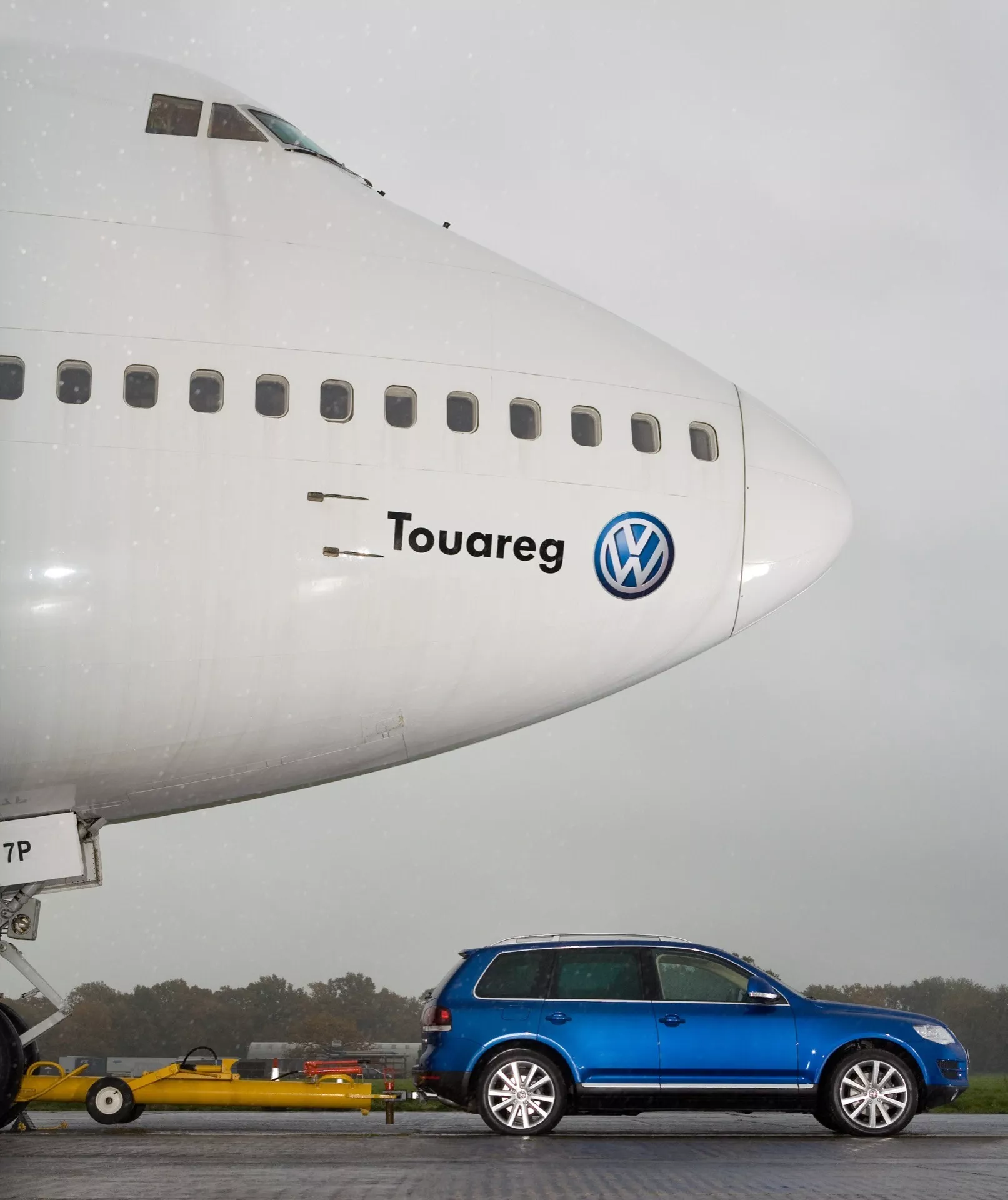 Volkswagen Touareg 2023 (III facelift) zaparkovaný kolmo před velkým obchodním letadlem v mlhavém prostředí, ukazující celý boční profil a designové linie vozu na pozadí srovnání velikosti inspirovaného letectvím, s viditelným logem a názvem modelu propojujícím SUV s letadlem