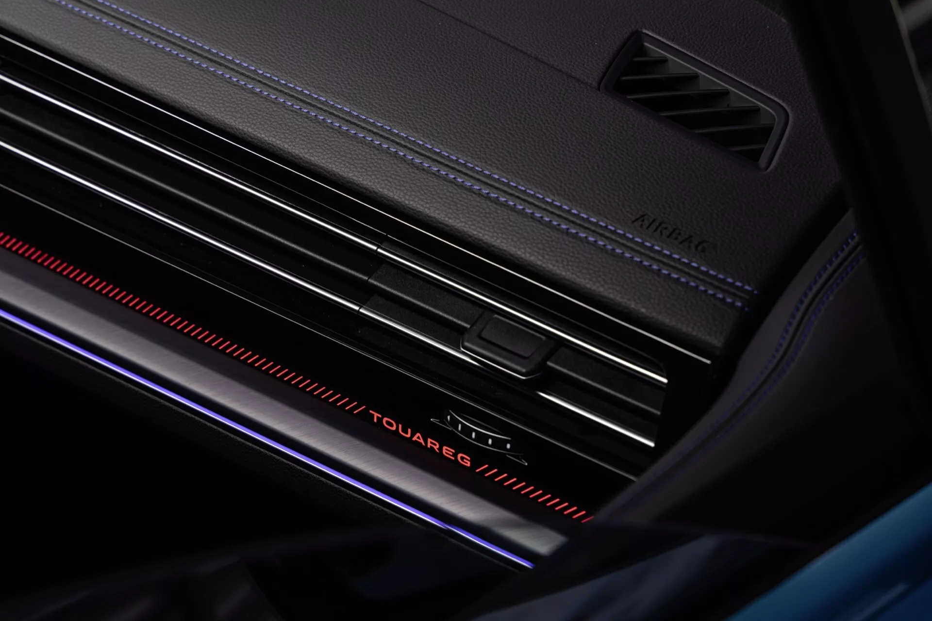 Detailní pohled na interiér Volkswagen Touareg facelift 2023 se zaměřením na design palubní desky, výraznou nápis 'Touareg' s ambientním osvětlením, ventilační mřížky se stříbrnými akcenty a atmosférický červený světelný pruh pro pocit luxusu