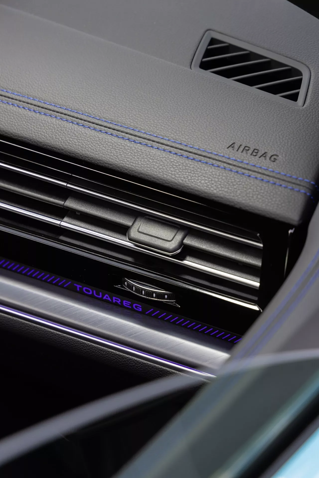 Detailní úhlový pohled na palubní desku na straně spolujezdce Volkswagen Touareg (III-Facelift) 2023 s označením AIRBAG, chromované výdechy klimatizace se středovým ovládacím knoflíkem, osvětleným nápisem 'TOUAREG', luxusními materiály se zdůrazněným prošíváním a rafinovanými prvky interiérového designu v prostředí se sníženou osvětleností zdůrazňujícími pokročilou interiérovou osvětlovací technologii a bezpečnostní prvky vozidla.