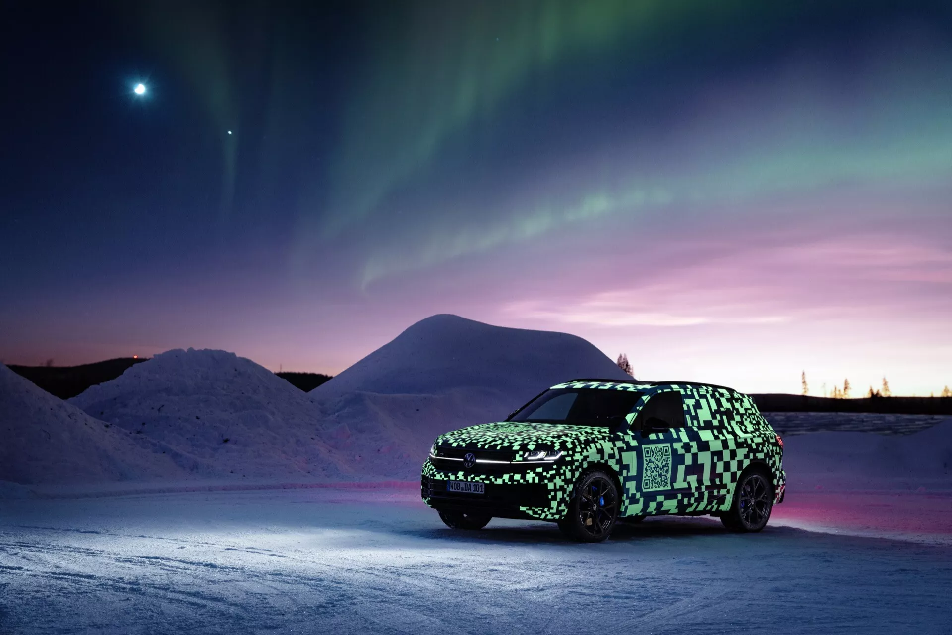 Prototyp VW Touareg (III-facelift) v maskovacím obalu na zasněženém povrchu pod polární září s jasným nebeským tělesem ve večerní obloze, zdůrazňuje pokročilé osvětlení a potenciální elektrickou nebo hybridní technologii.