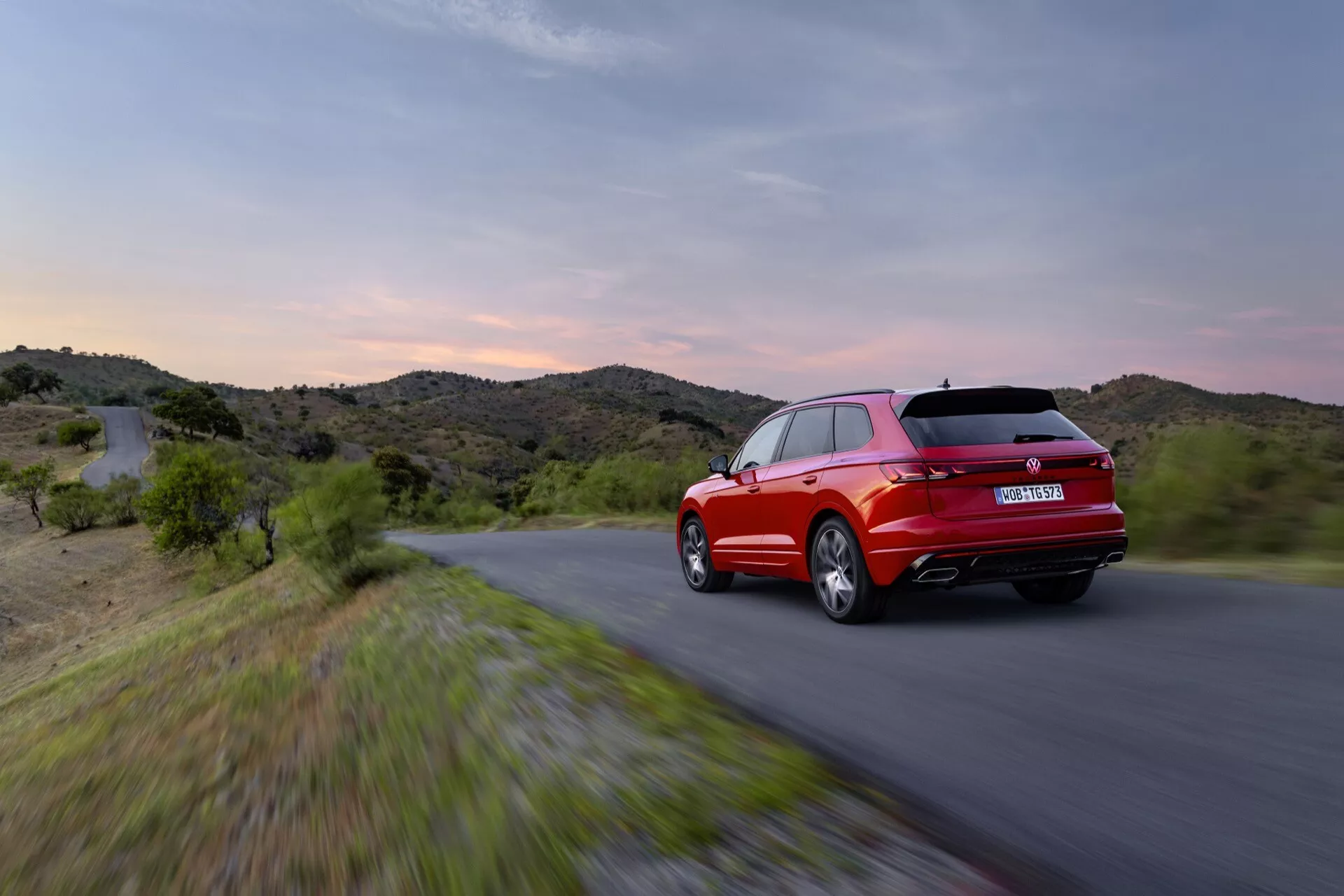 VW Touareg III Facelift 2023 v pohybu z pohledu ze zadního tříčtvrtečního úhlu s LED zadními světly a sportovní konturou proti pozadí večerní oblohy a kopcovité krajiny, zdůrazňující jeho červenou barvu, aerodynamický design a výkonnostní prvky.
