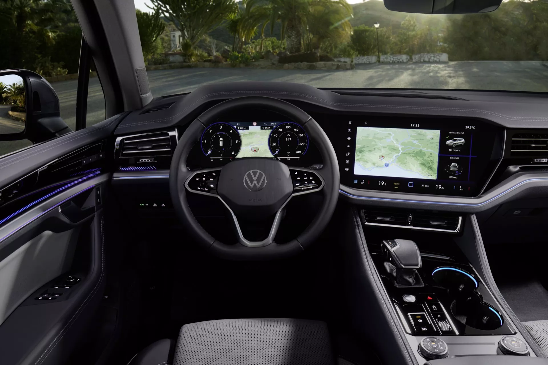 Interiér Volkswagen Touareg III facelift 2023 z pohledu zadních sedaček směrem k digitálnímu palubnímu desce s přirozeným denním osvětlením, představující multifunkční volant s logem, digitální přístrojový panel, velký dotykový infotainment systém s GPS, elegantní dotykové ovládání klimatizace, minimalistickou páku automatické převodovky a centrální konzolu s ambientním osvětlením, vysoce kvalitním provedením a bezpečnostními prvky