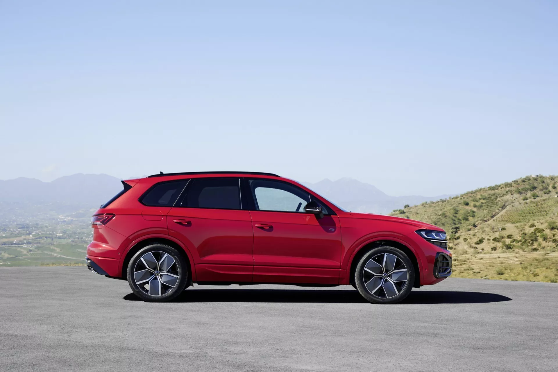 Model VW Touareg III po faceliftu v červené barvě, zaparkovaný na rovném povrchu s horskou krajinou a jasným nebem na pozadí, ukazující svůj boční profil s detailním pohledem na slitinová kola, kliky dveří, boční zrcátka s integrovanými směrovými signály, střešní ližiny, sportovně střiženou siluetu ve stylu kupé a tónovaná okna.