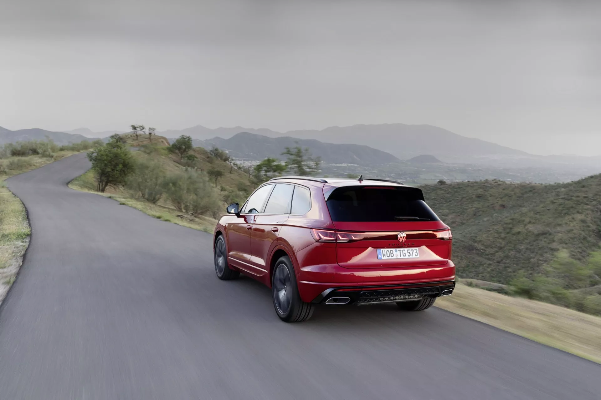 Dynamický zadní pohled na VW Touareg 2023 na horské silnici, předvádějící aerodynamický design, dvojité výfuky a sportovní provedení ve výrazné červené barvě