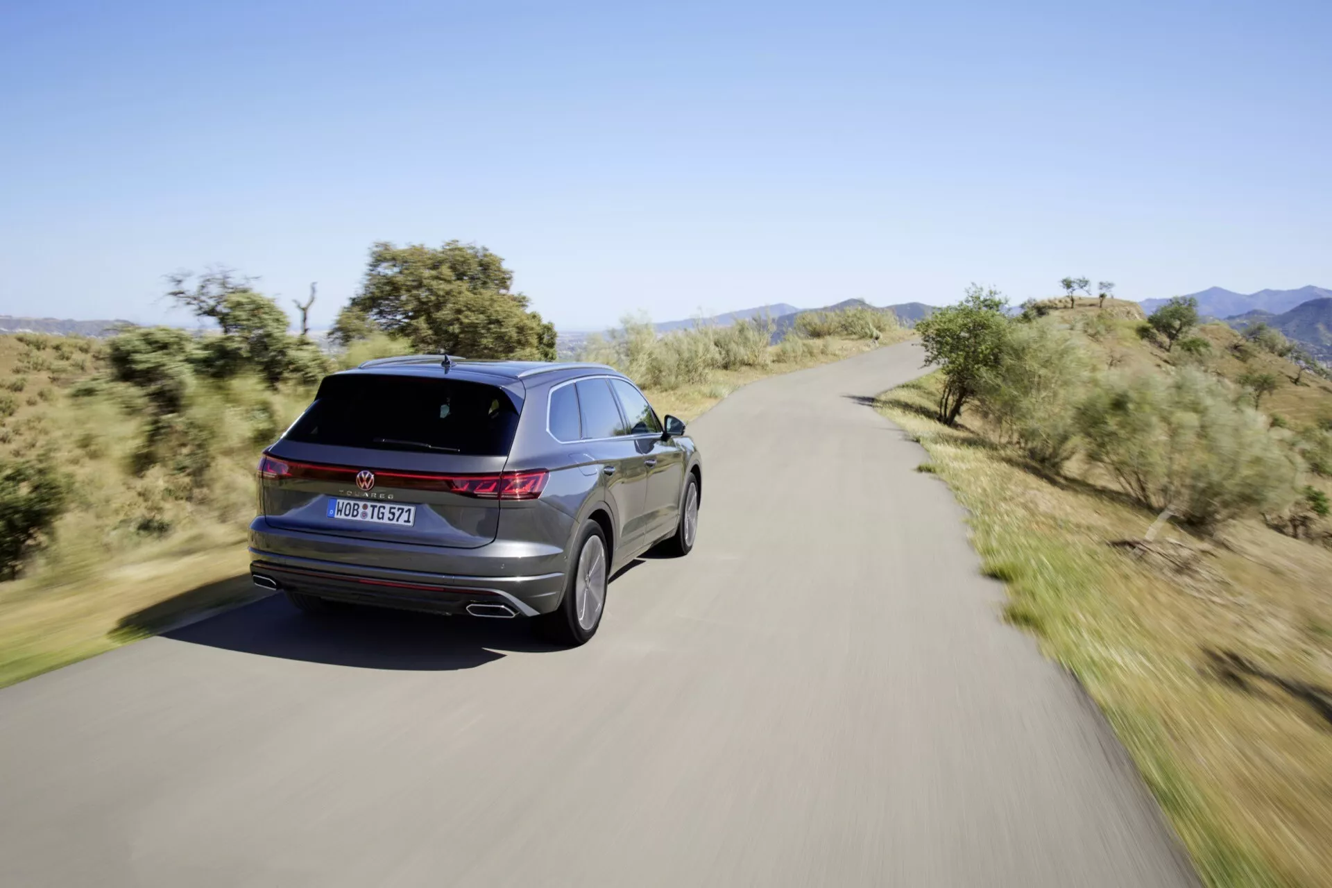 Zadní tříčtvrteční pohled na VW Touareg III-Facelift 2023 na scénické silnici, ukazuje LED zadní světla, charakteristické označení, dvojité výfukové špunty, slitinová kola, elegantní linii střechy a lesklý lak.