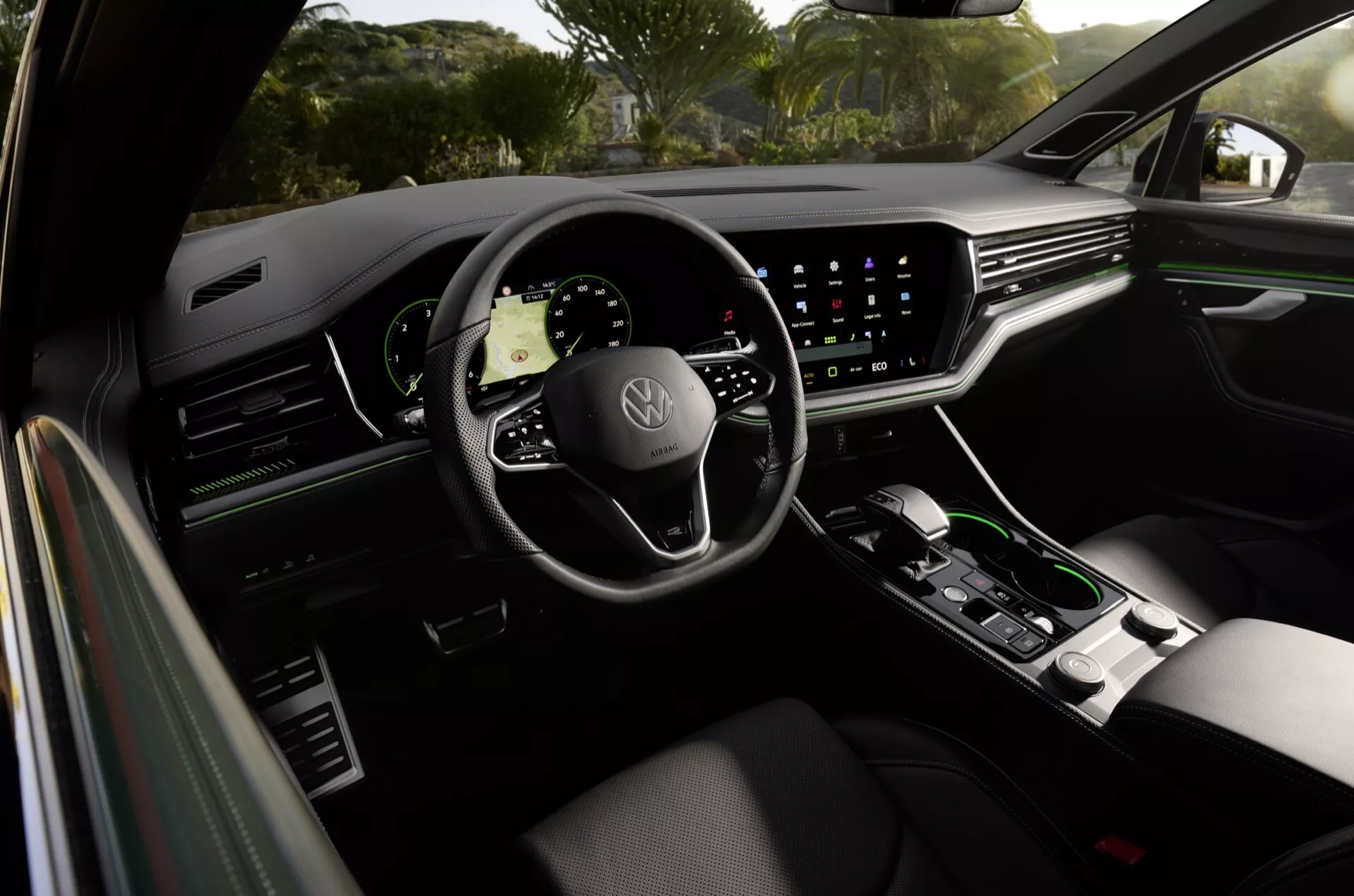 Interiér Volkswagen Touareg 2023 z pohledu ze zadního sedadla, zobrazující panel dveří řidiče, prémiové ergonomické sedadlo řidiče, volant se znakem Volkswagen s ovládacími prvky, digitální přístrojový panel a velký dotykový informačně-zábavní systém s navigací, kromě minimalistických ovládacích prvků klimatizace a selektoru automatické převodovky na středové konzole, což odráží luxusní vlastnosti a pokročilou technologii