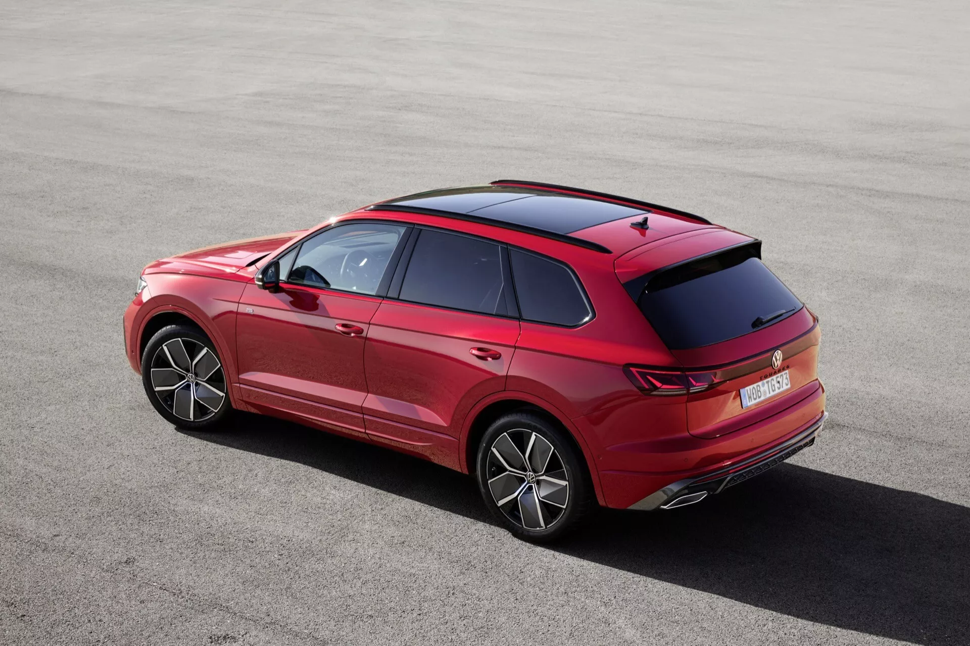 VW Touareg III po faceliftu z roku 2023 viděn z vysokého úhlu zezadu a ze strany, předvádějící jeho elegantní siluetu, výrazné linie karoserie, linii střechy, detailní design zadního spoileru a anten, inovativní tvar a design zadních světel a nárazníku, jasně viditelná kola s stylovými ráfky, zadní čelní sklo, prvky designu zadních dveří s kličkami a lištami, s náznaky předních dveří a bočních zrcátek, představující technologické prvky jako jsou zadní parkovací senzory, vysoko umístěné brzdové světlo, technologie LED nebo OLED zadních světel, a možná aerodynamická přídavky na zadním nárazníku, zdůrazňující proporce vozidla a jeho postoj.