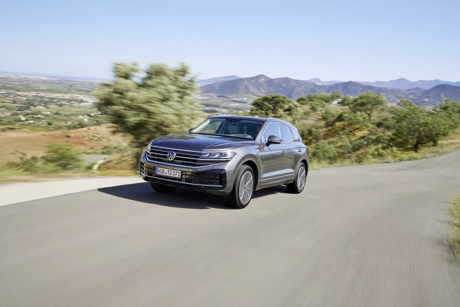 Model VW Touareg 2023 po faceliftu dynamicky jedoucí po zpevněné venkovské silnici, prezentuje svůj moderní design a výkonnostní prvky, včetně LED světlometů, luxusního metalického laku a elegantních slitinových kol, na pozadí horské krajiny