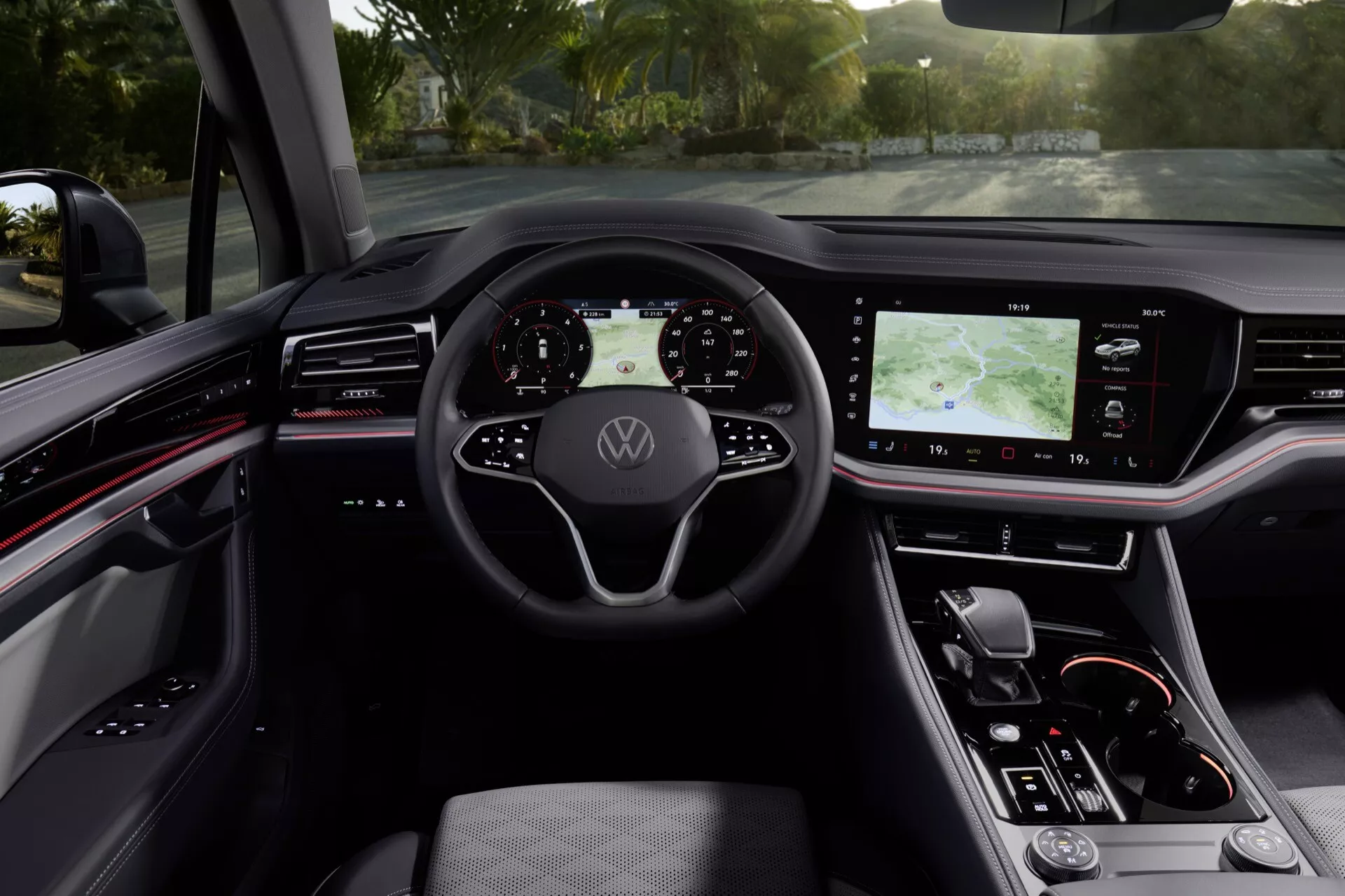 Interiér VW Touareg 2023 zobrazující volant s logem Volkswagen, digitální přístrojovou desku, velký dotykový informačně-zábavní systém na středové konzole, ergonomické sedadlo řidiče potažené kůží a moderní designové prvky s červenými akcenty z pohledu řidiče.