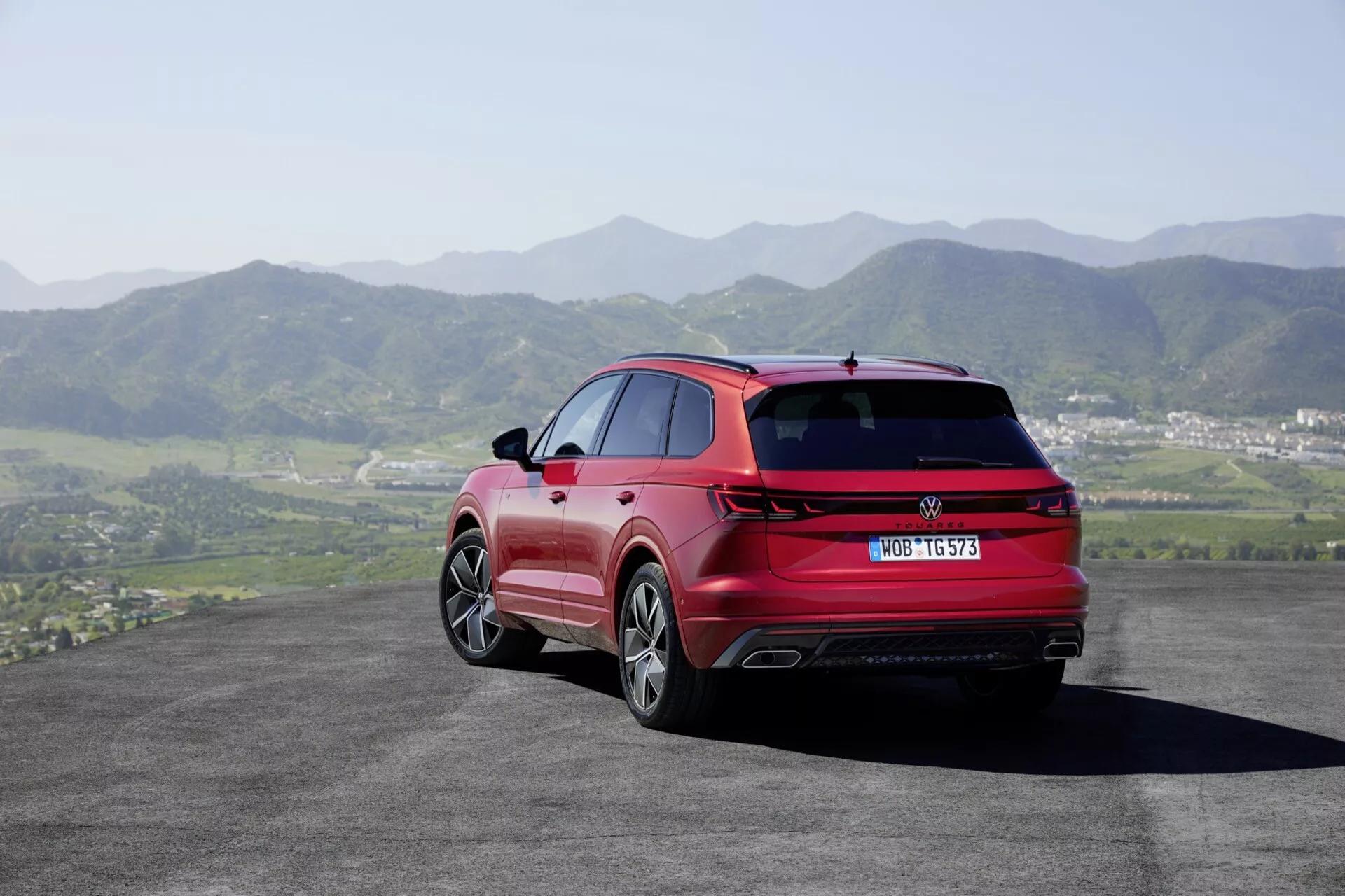 VW Touareg III Facelift 2023 v červené barvě - pohled zezadu a z boku ukazuje LED zadní světla, dvojité výfuky, slitinová kola a stříbrné střešní ližiny na pozadí válcujících se kopců a jasné oblohy