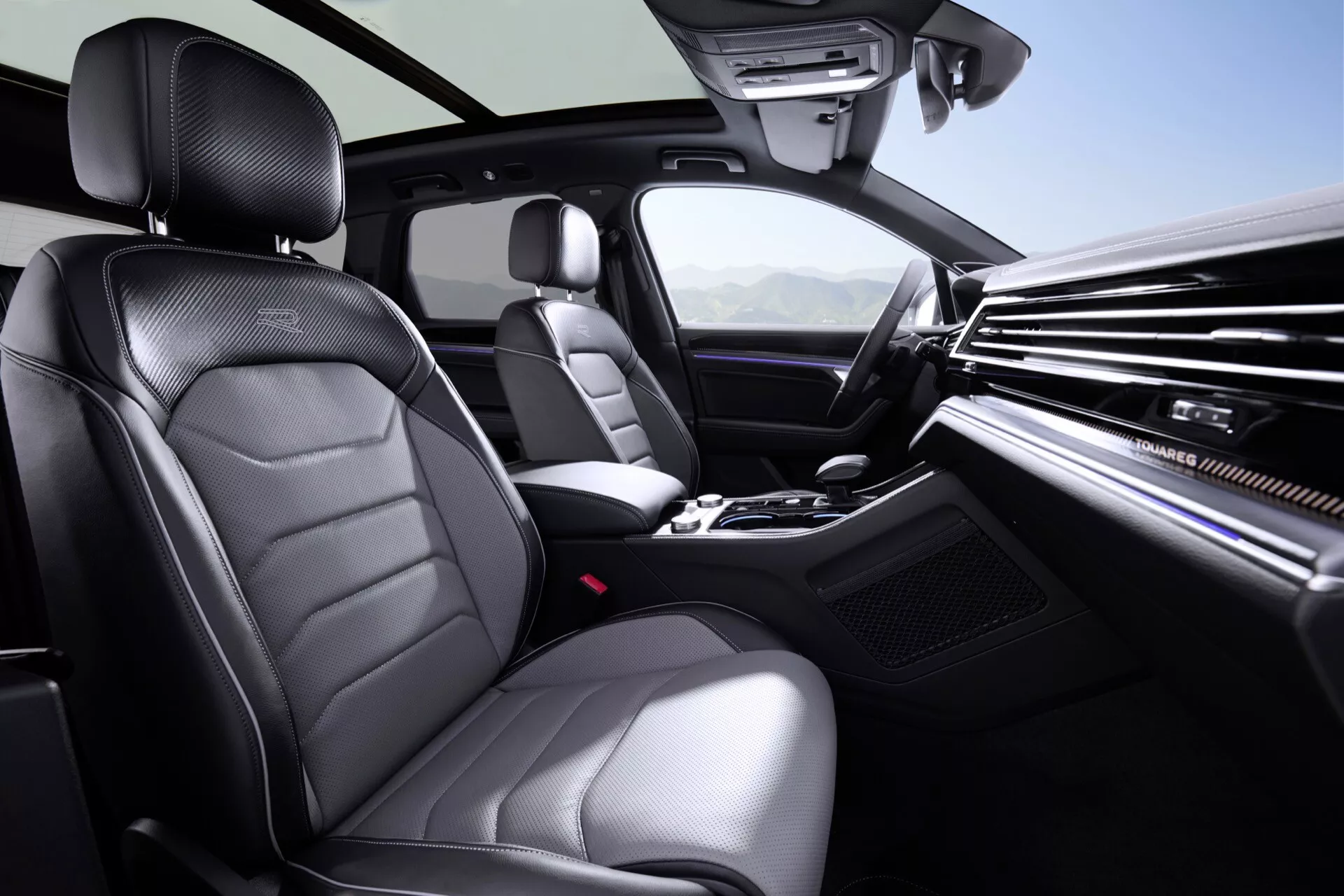 Interiér VW Touareg 2023 prezentující luxusní a technologické prvky jako jsou sedadla z kvalitní kůže, palubní deska s dřevěným obložením, páka automatické převodovky, klimatizace s dvojí zónou, multifunkční volant, ambientní osvětlení panoramatické střechy a konzole informačně-zábavního systému.