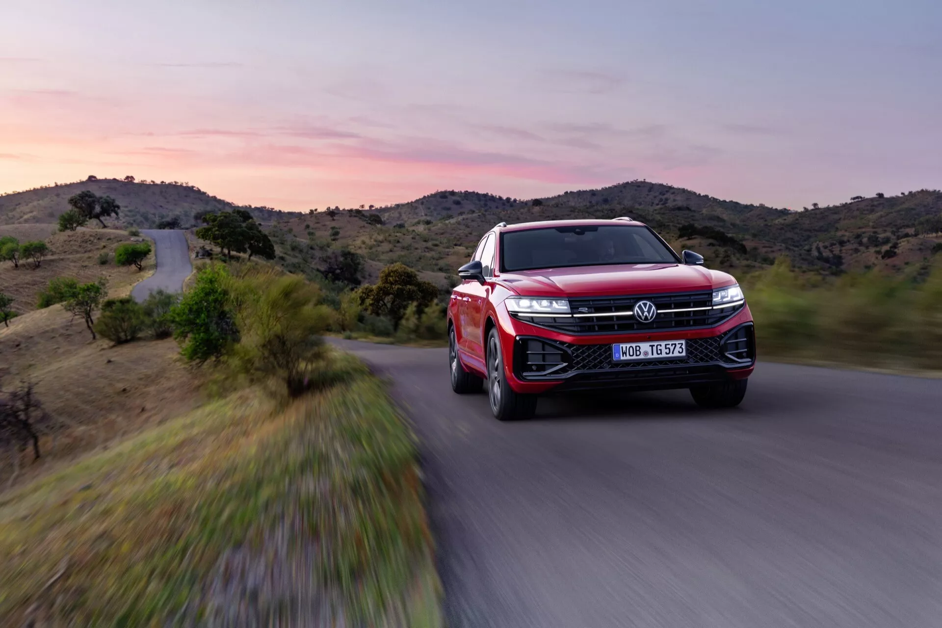 VW Touareg III-facelift 2023 v červené barvě, zobrazující pohled z předního tříčtvrtečního úhlu s důrazem na LED světlomety, velkou mřížku s emblémem, sportovně aerodynamický design, pokročilé slitinové kola a funkce ADAS v dynamickém pohybu na scénické silnici za soumraku