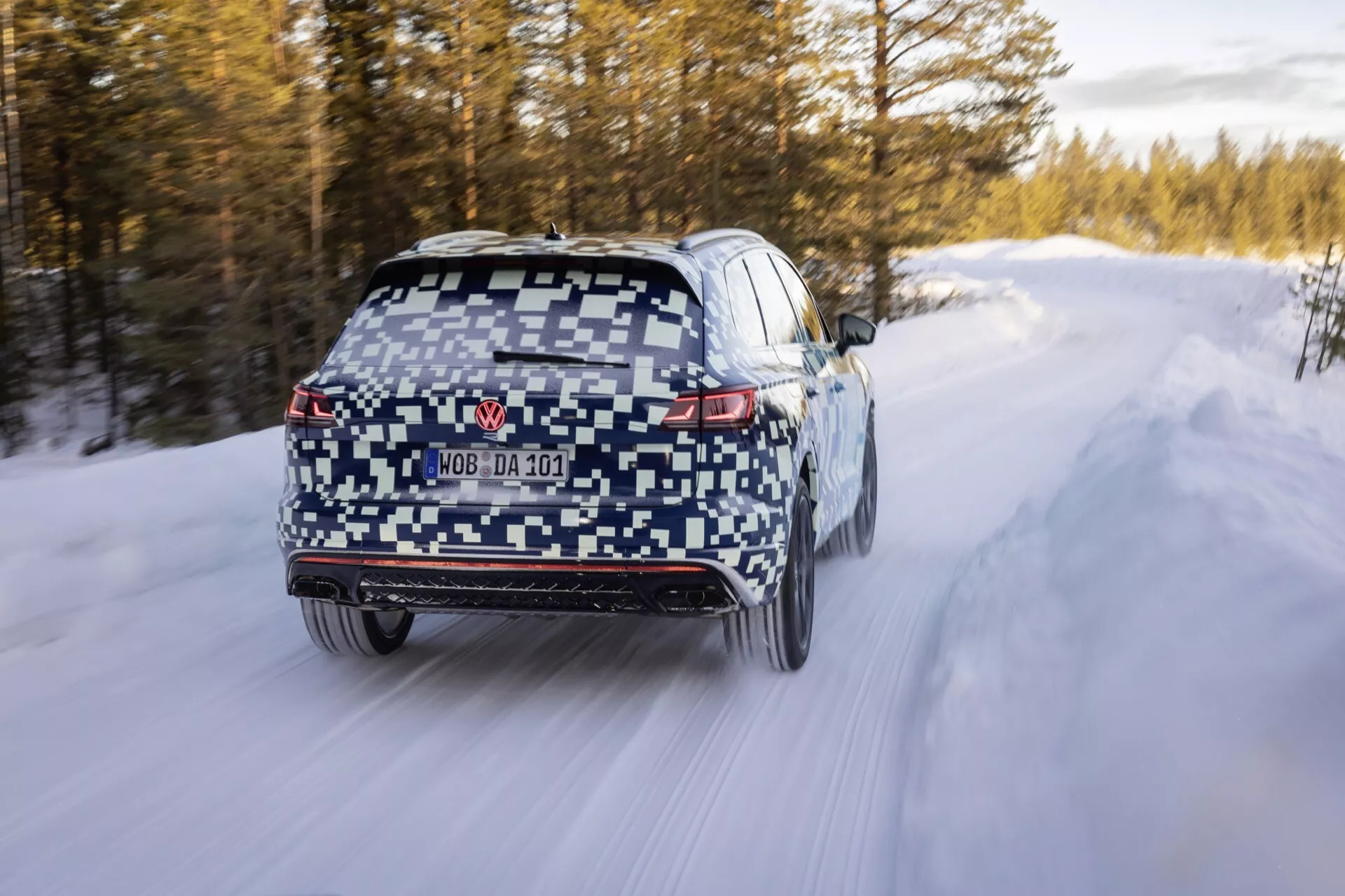 VW Touareg III Facelift 2023 v maskování jedoucí po zasněžené silnici s LED zadními světly, zadním difuzorem, stěračem zadního okna, částečně skrytou SPZ, dvojitými výfuky, senzory na zadním nárazníku, rozmazaním pohybu naznačující pohyb, zimními pneumatikami a potenciálním pohonem všech kol