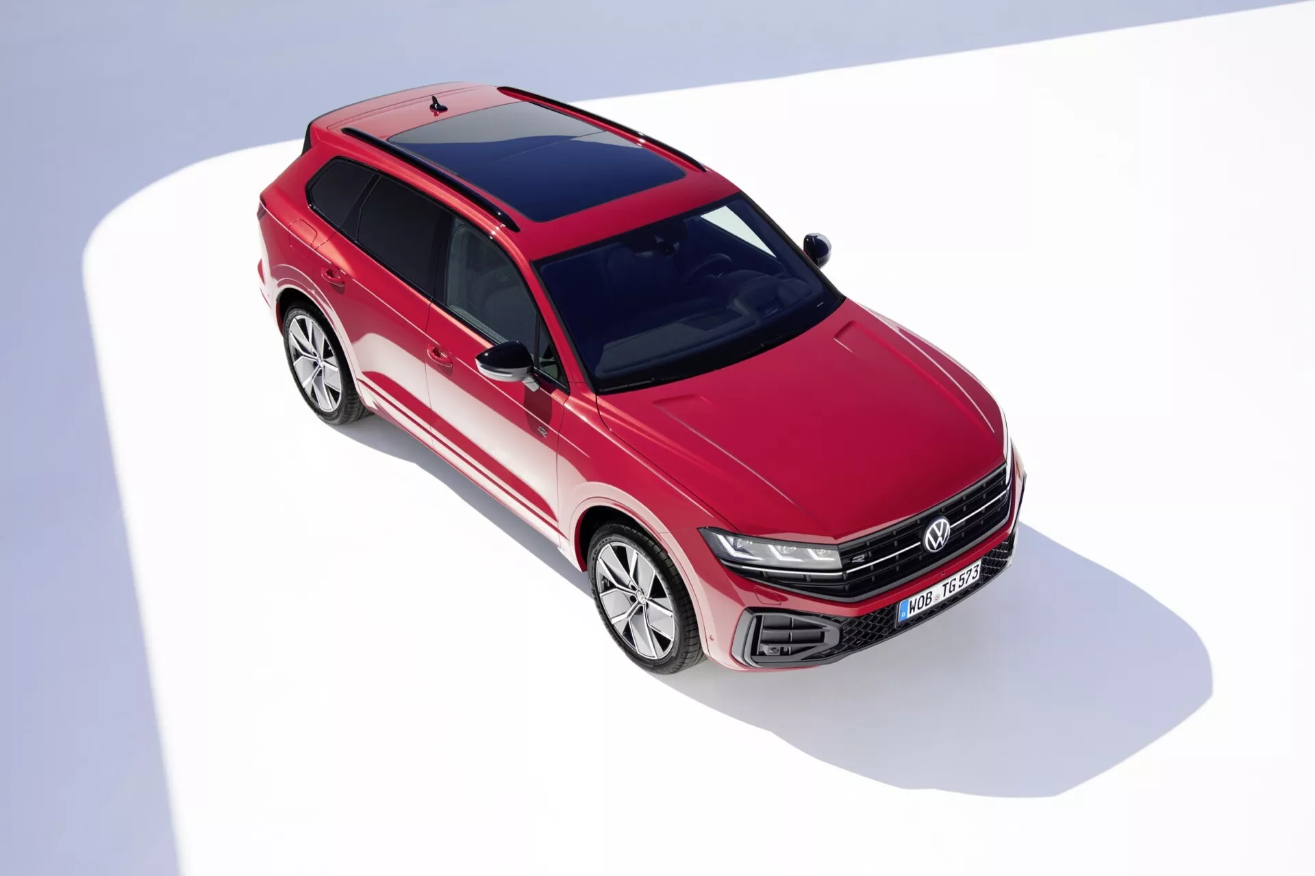 Pohled z vysokého úhlu na přední část a stranu vozu 2023 VW Touareg III Facelift v červené barvě, zobrazující designové prvky včetně mřížky s horizontálními lamelami a emblémem, LED předních světel, aerodynamicky tvarovaného předního nárazníku s otvory pro přívod vzduchu, profilovaných linií kapoty, rozsáhlého čelního skla, slitinových kol s vícepaprskovým designem, panoramatické střechy a aerodynamických zpětných zrcátek na bílém pozadí