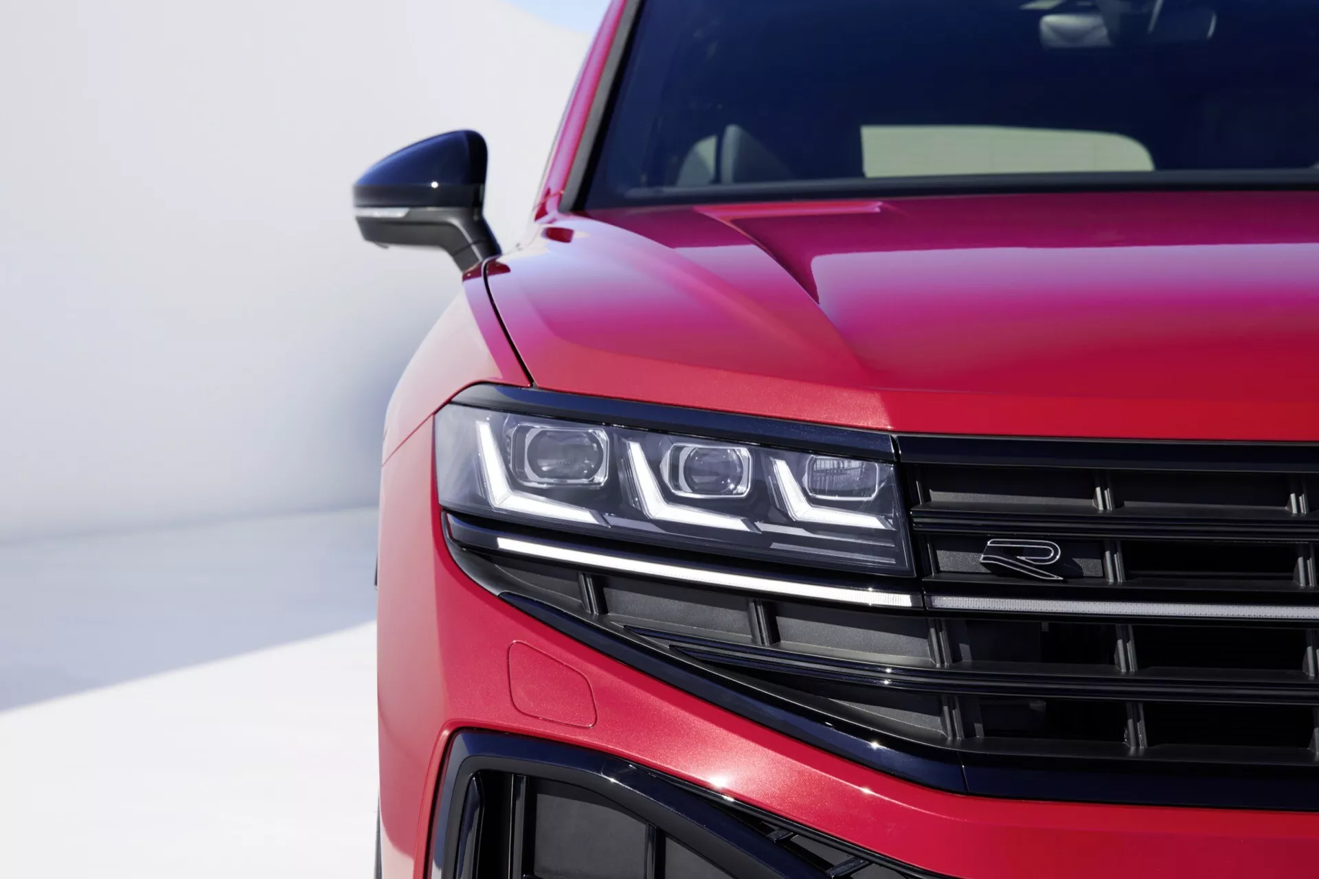 VW Touareg III-Facelift rok 2023 s lesklým metalickým červeným lakem s detailním pohledem na přední levý čtvrtletní pohled ukazující sestavu LED světlometů, část mřížky s horizontálními lamelami, podrobný pohled na přední nárazník a aerodynamické linie na kapotě proti rozmazanému pozadí zdůrazňujícím moderní design a technologické vlastnosti vozu.