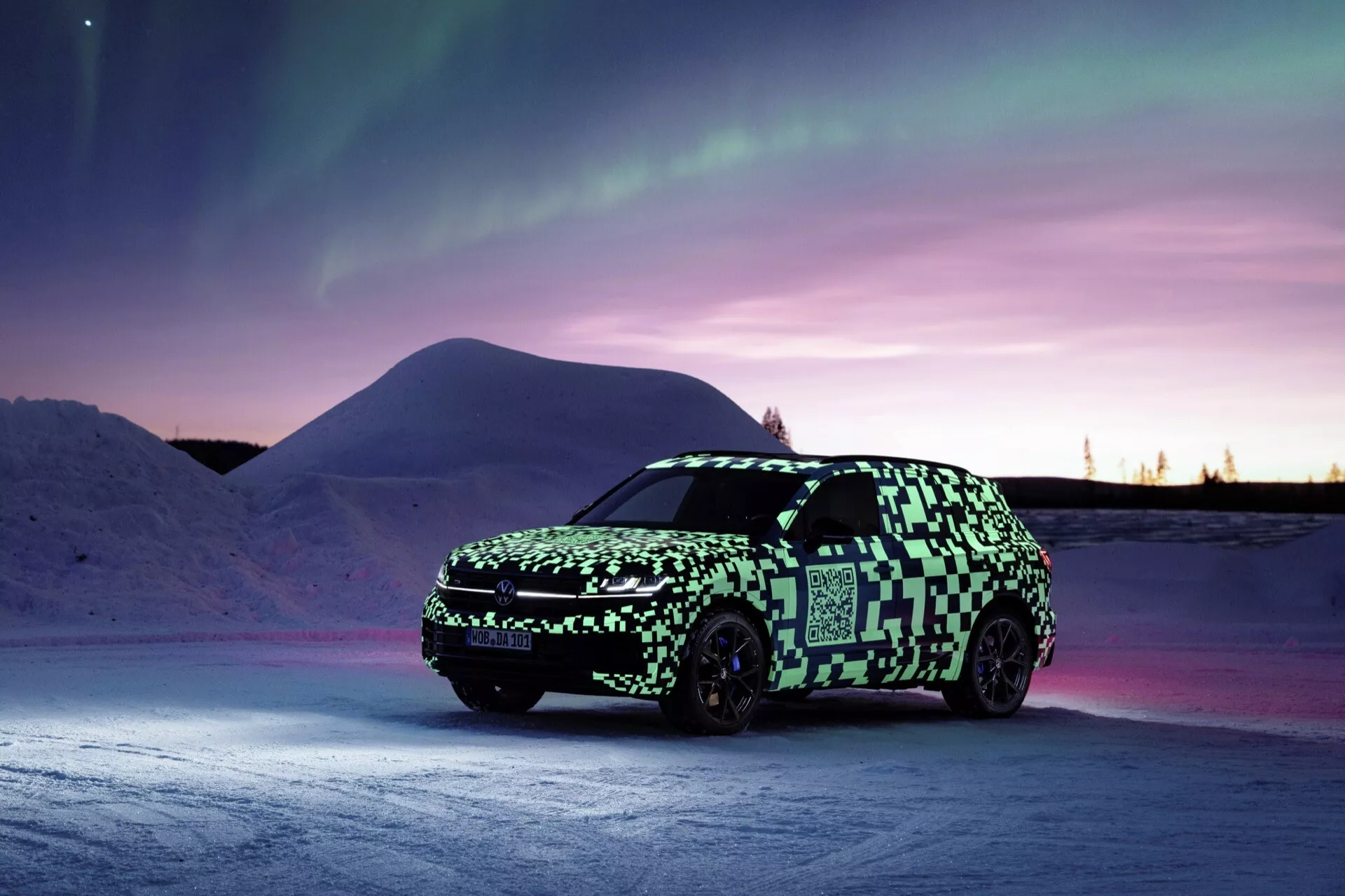 Model Volkswagen Touareg III facelift 2023 představen v zimním soumraku, zobrazení z perspektivy tři čtvrtiny zepředu s důrazem na LED světlomety, přední mřížku a maskovací obal na pozadí polární záře a zasněžené krajiny