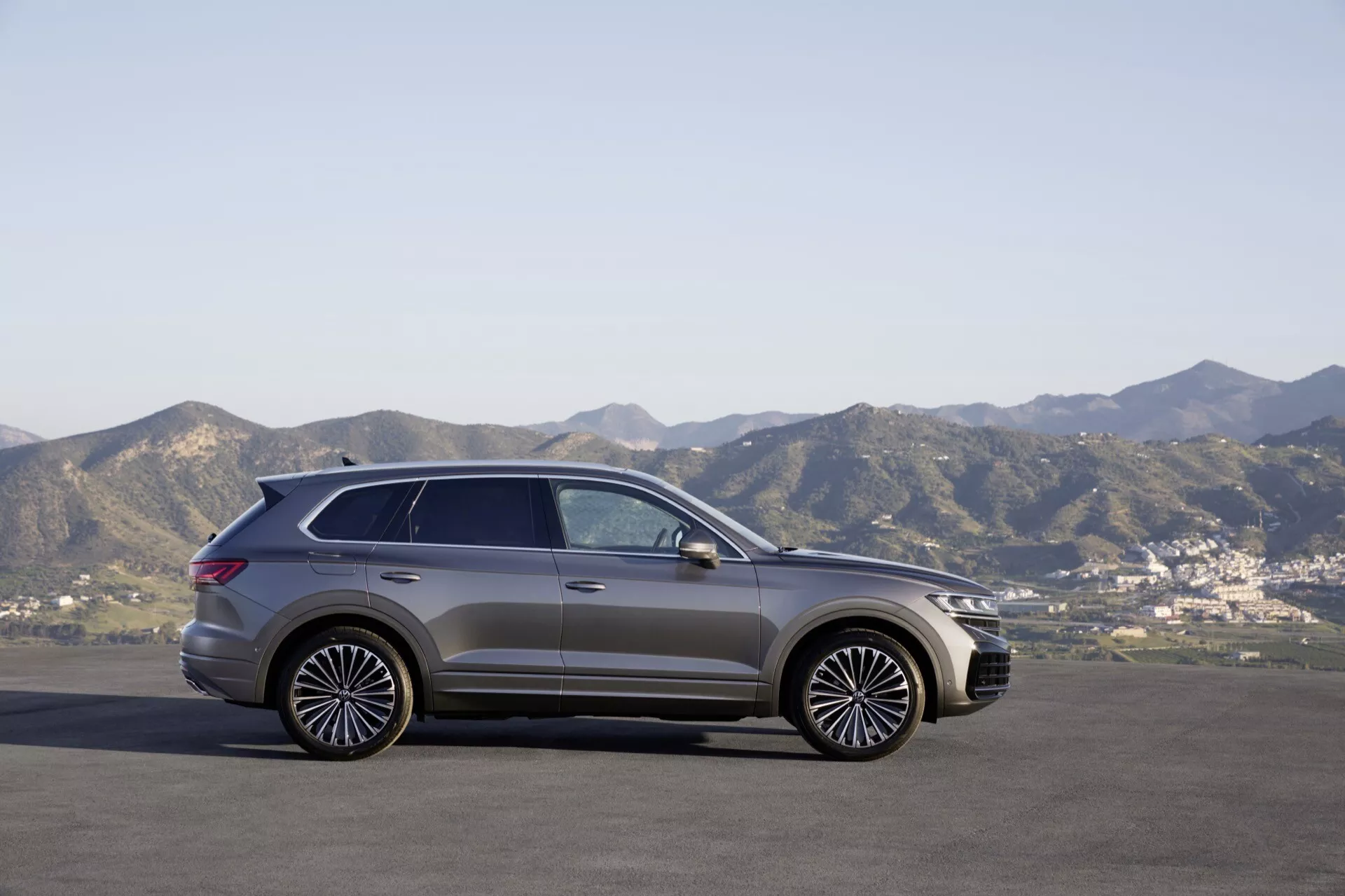 Model VW Touareg III po faceliftu v roce 2023 v pohledu z boku zaparkovaný na rovném povrchu proti horské kulisě pod jasnou oblohou, ukazující jeho moderní design SUV, stylové slitinové kola a panoramatickou střechu.