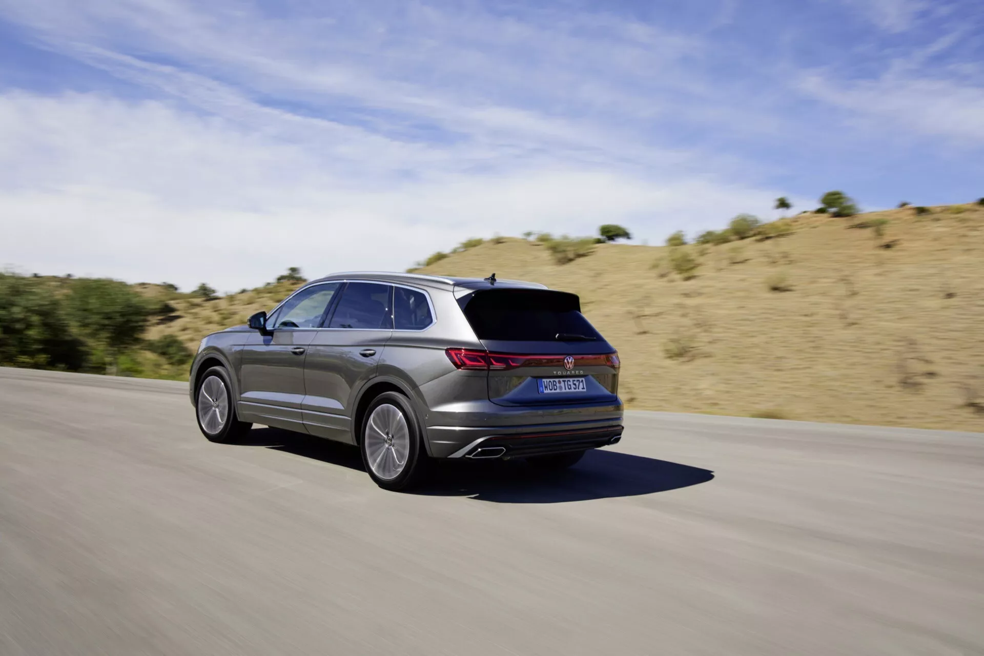 VW Touareg III facelift 2023 v pohybu na asfaltové silnici, zachycený ze zadního tříčtvrtečního úhlu ukazuje jeho elegantní design, pětipaprsková slitinová kola, metalický finiš, LED zadní světla, integrované výfuky a sportovní estetiku na pozadí suché, kopcovité krajiny pod jasně slunečnou oblohou