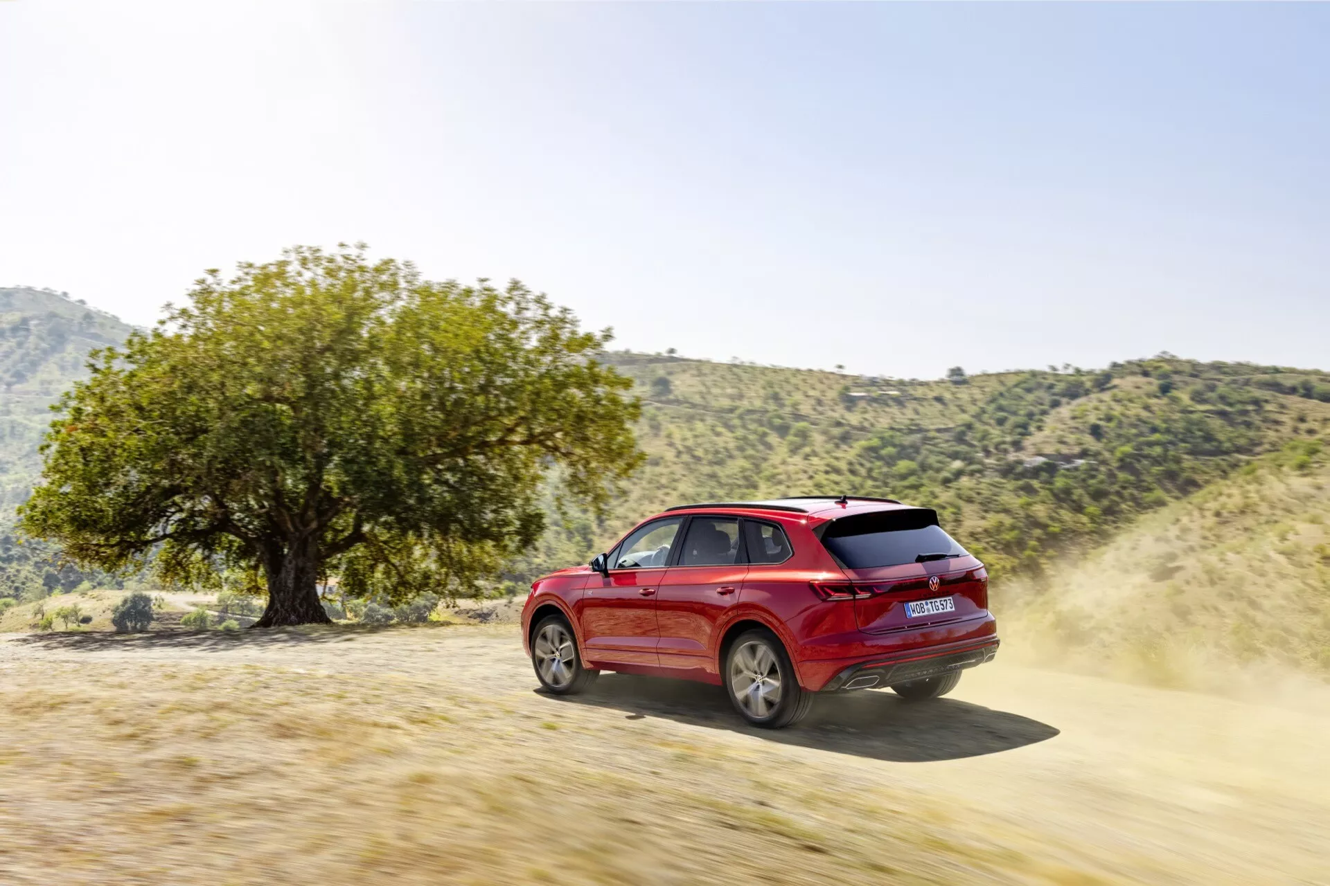 Červené SUV Volkswagen Touareg III Facelift 2023 předvádějící schopnosti v terénu na nezpevněné cestě, záběr ze tříčtvrtečního zadního úhlu zdůrazňující jeho dynamický design, ráfky kol, boční dveře, boční a zadní okna, s prachem naznačujícím pohyb a výkonnostní charakteristiky jako výkon motoru a pohon 4x4, v přírodním venkovním prostředí naznačujícím dobrodružství a všestrannost v terénu.