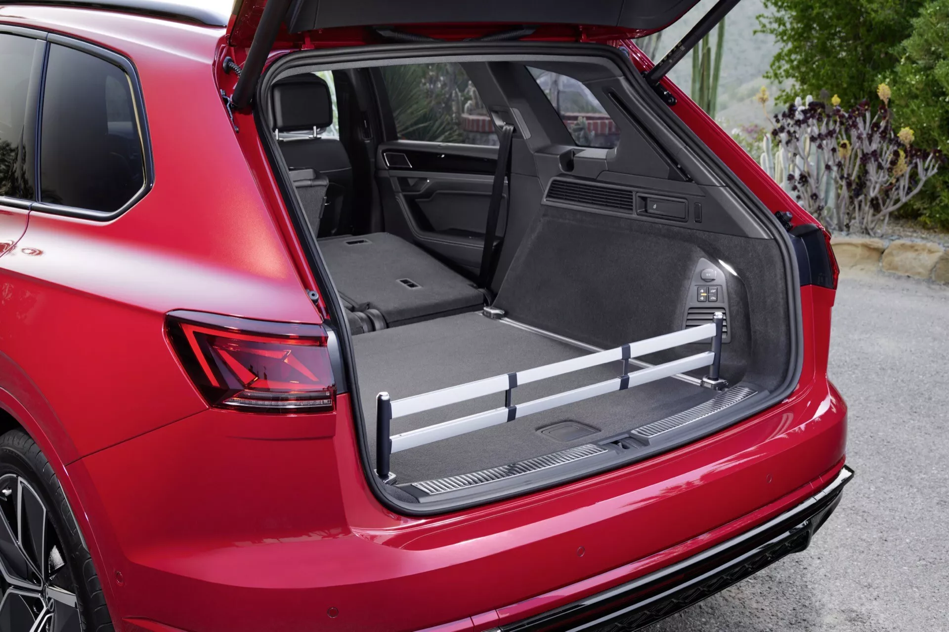 Detailní snímek z roku 2023 Volkswagen Touareg III po faceliftu ukazující prostorný nákladní prostor s plochou nákladovou plochou, ochranným obložením a integrovanými úložnými přihrádkami z pohledu ze zadní pravé části pod úhlem tři čtvrtiny, s 12V zásuvkou, nastavitelnými nákladními lištami a částečně sklopenými zadními sedadly pro rozšíření prostoru, zvýrazněno červeným exteriérem vozidla a sportovním zadním spoilerem.